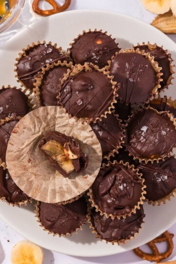 A stack of frozen banana bites with chocolate coating, pretzels, and peanuts, next to a single bite showing the banana and caramel center inside.