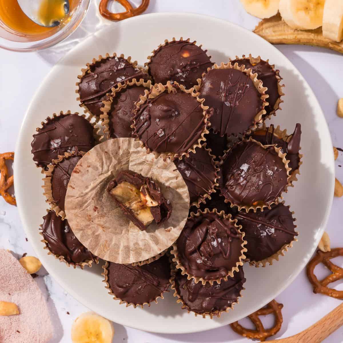 A stack of frozen banana bites with chocolate coating, pretzels, and peanuts, next to a single bite showing the banana and caramel center inside.