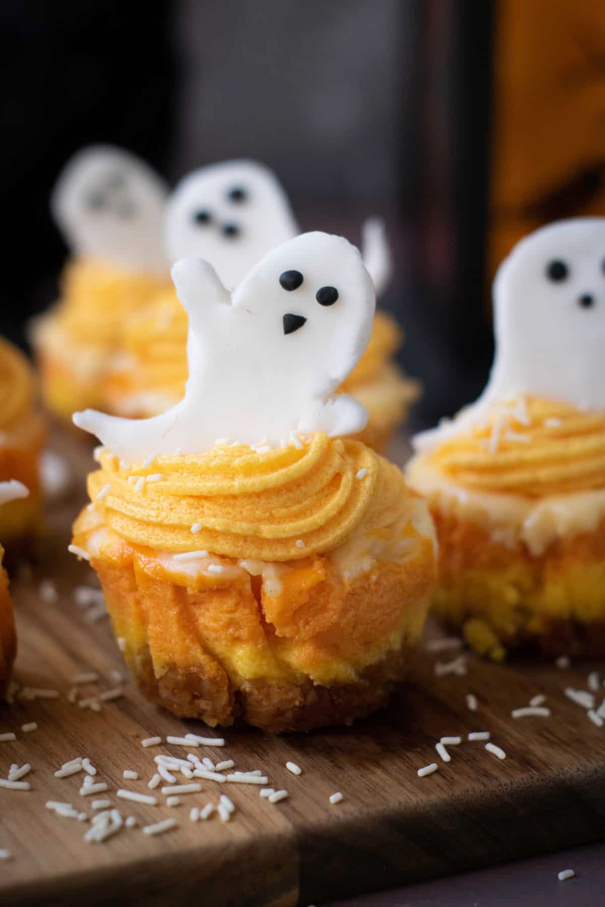 Mini Halloween cheesecakes with orange frosting and ghost toppers served on a wooden board.