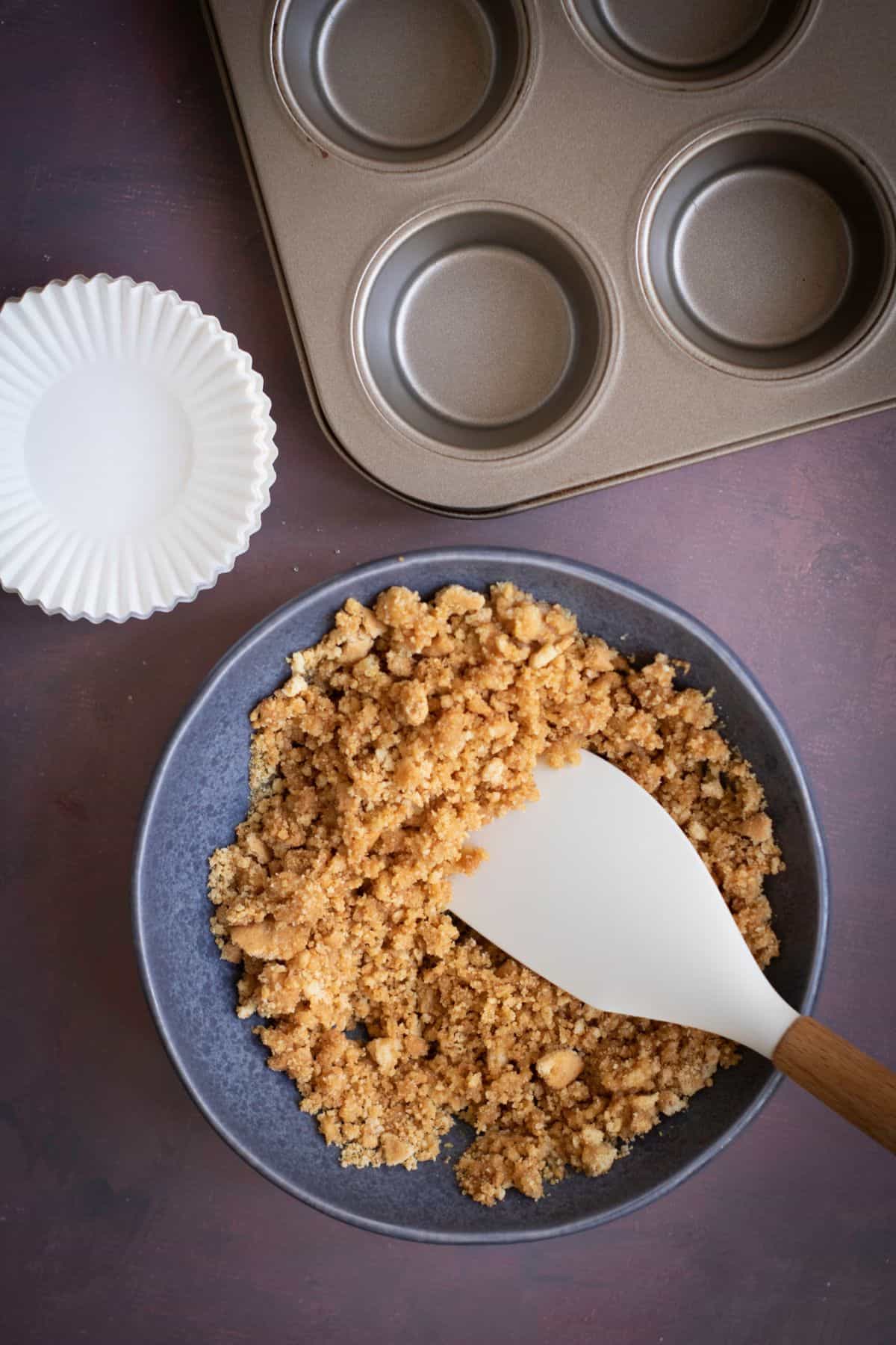 Graham cracker crust mixture in a large bowl with a muffin tin beside it.