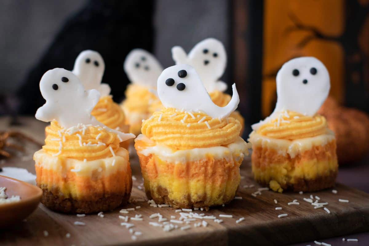 Mini Halloween cheesecakes with orange frosting and ghost toppers served on a wooden board.