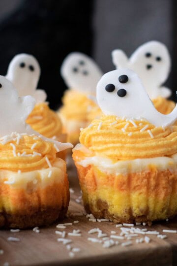 Mini Halloween cheesecakes with orange frosting and ghost toppers served on a wooden board.