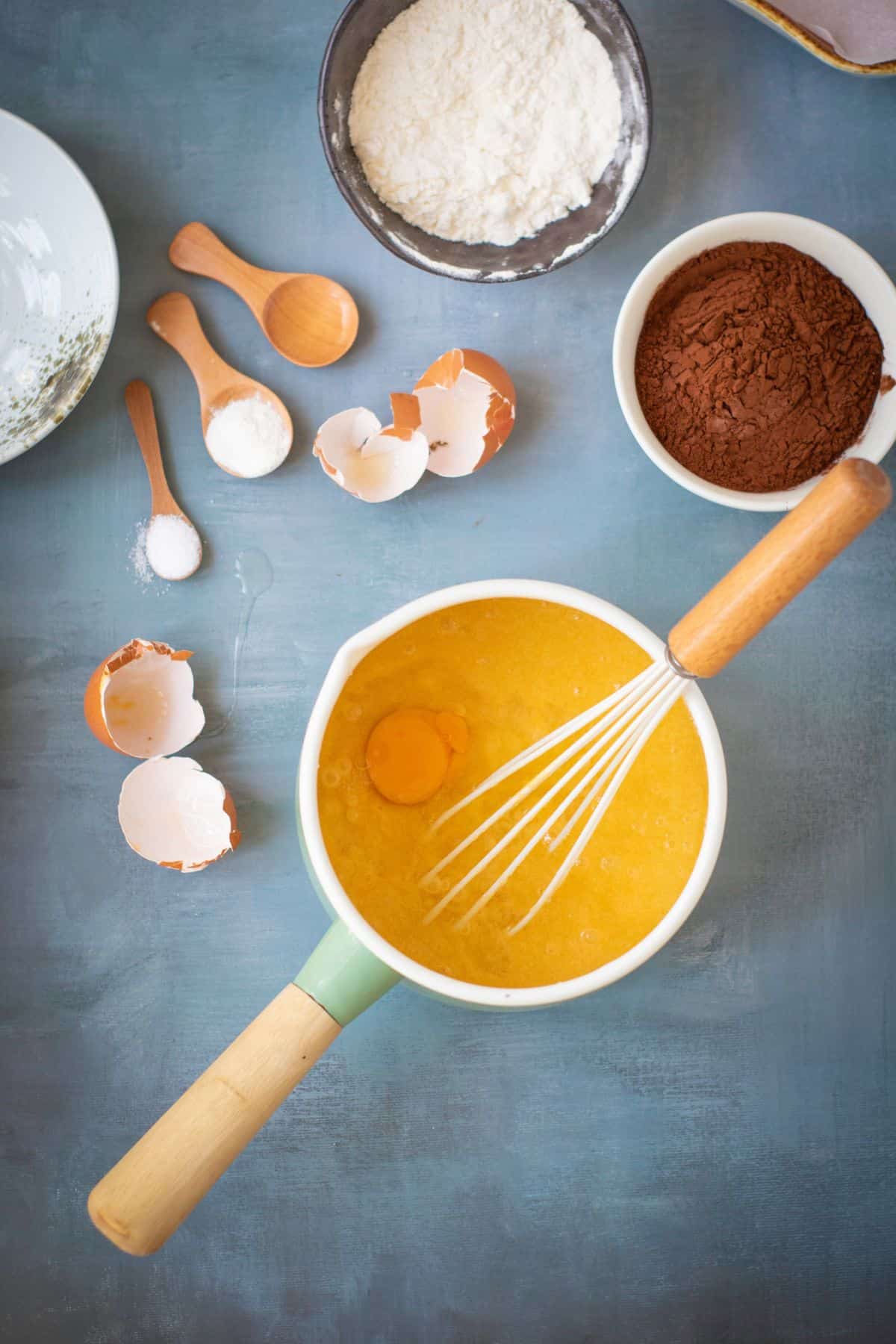 An egg being whisked in a mixing bowl using a whisk with tools and other ingredients nearby.