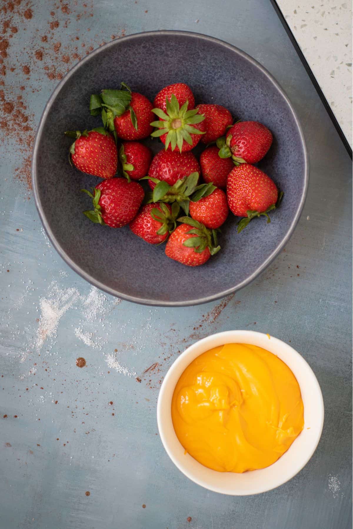 A fresh strawberries in a bowl next to a small dish of melted orange candy coating.