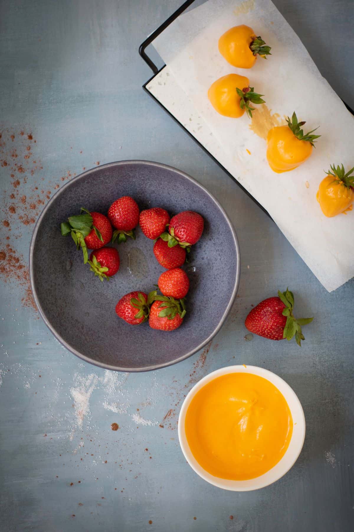 A strawberry being dipped into melted orange candy coating and placed on parchment paper, with other uncoated strawberries in a medium bowl nearby.