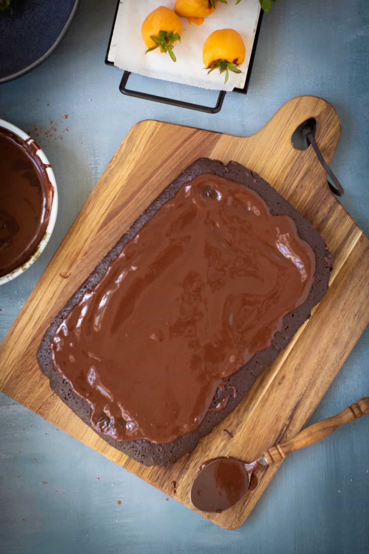A chocolate brownie cake being placed on a rustic wooden board, topped with a smooth layer of chocolate ganache.