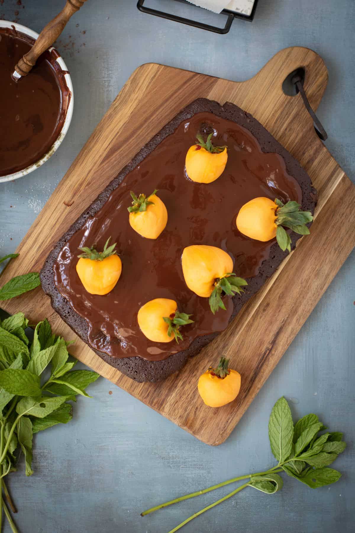 Chocolate brownie cake on a rustic wooden board, spread with ganache and being topped with orange candy-coated strawberries.