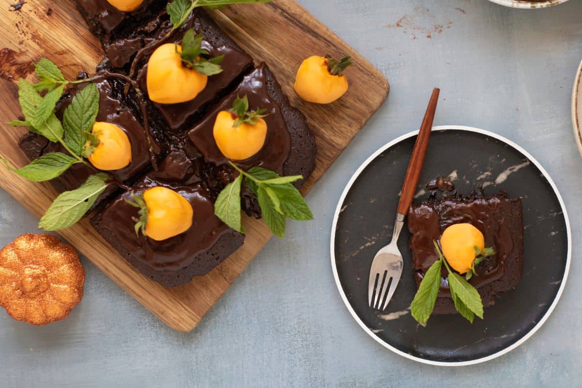 Chocolate pumpkin patch brownie cake being garnished with candy-coated strawberries and mint leaves, served on a rustic wooden board and placed a single cake nearby.