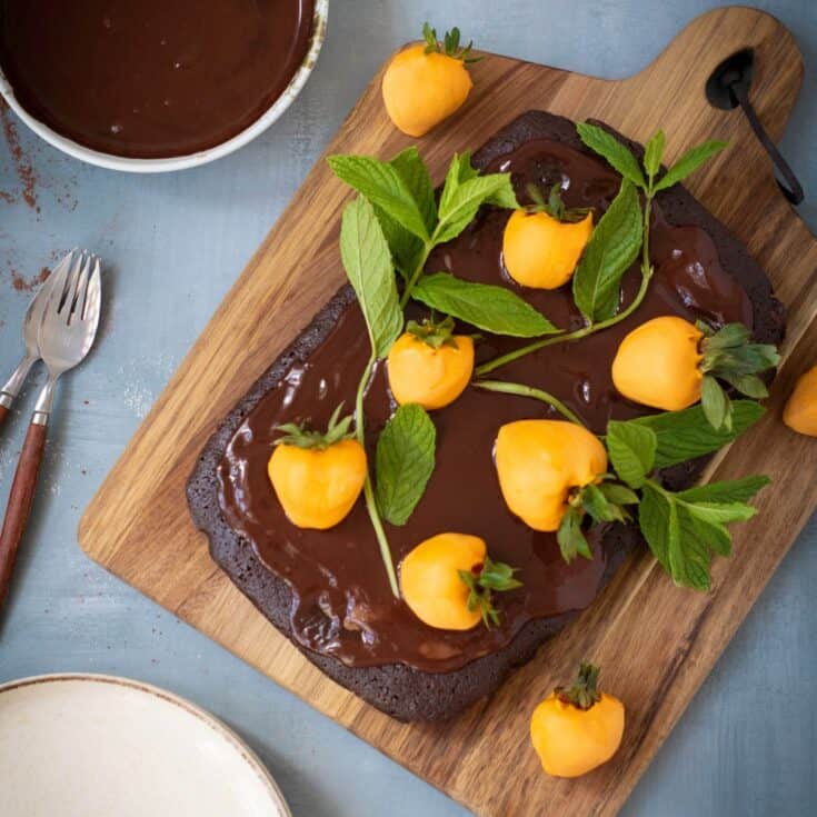 Chocolate pumpkin patch brownie cake being garnished with candy-coated strawberries and mint leaves, served on a rustic wooden board.