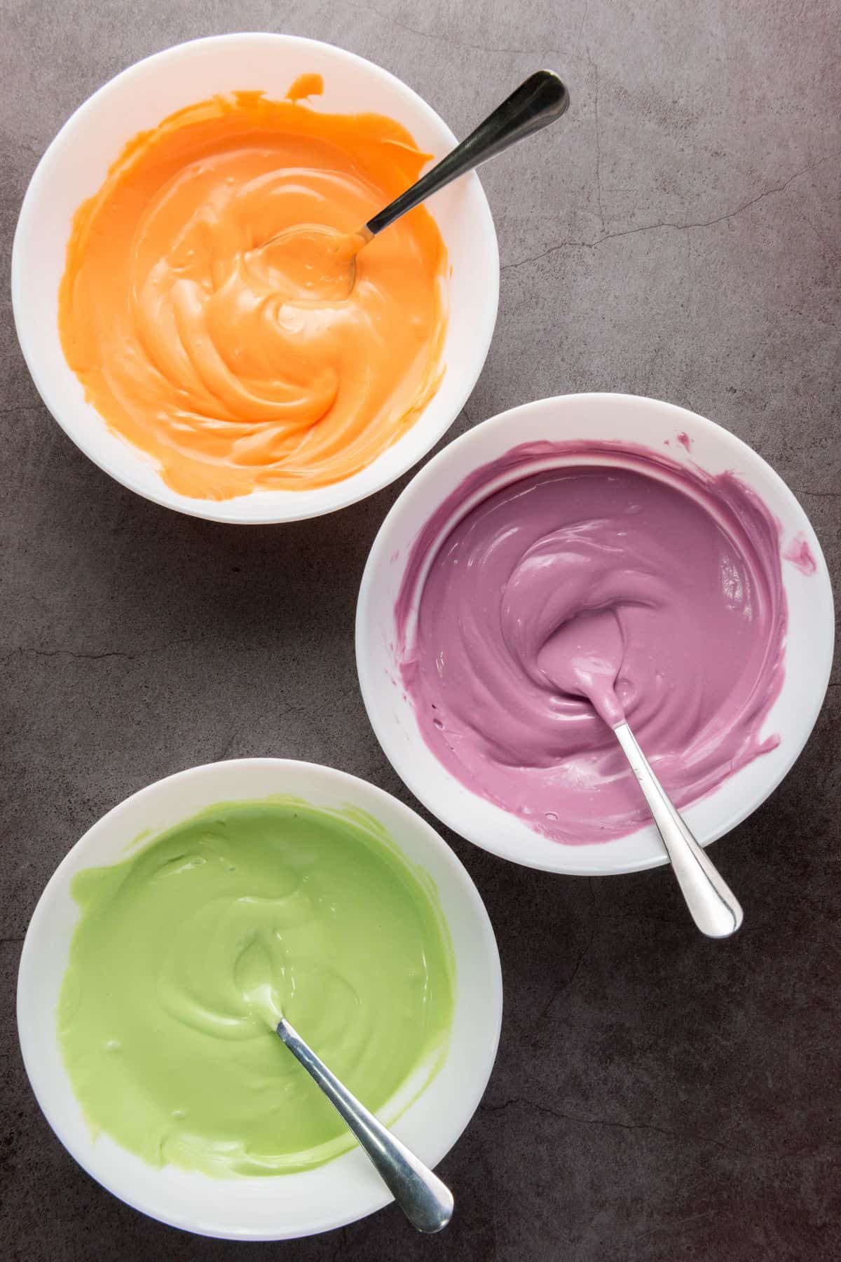 Three bowls filled with melted purple, green, and orange candy melts, ready for dipping Halloween cookies.