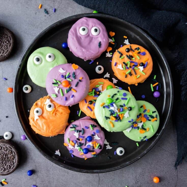 A plate full of colorful Halloween-dipped cookies surrounded by extra plain Oreos, ready to serve.
