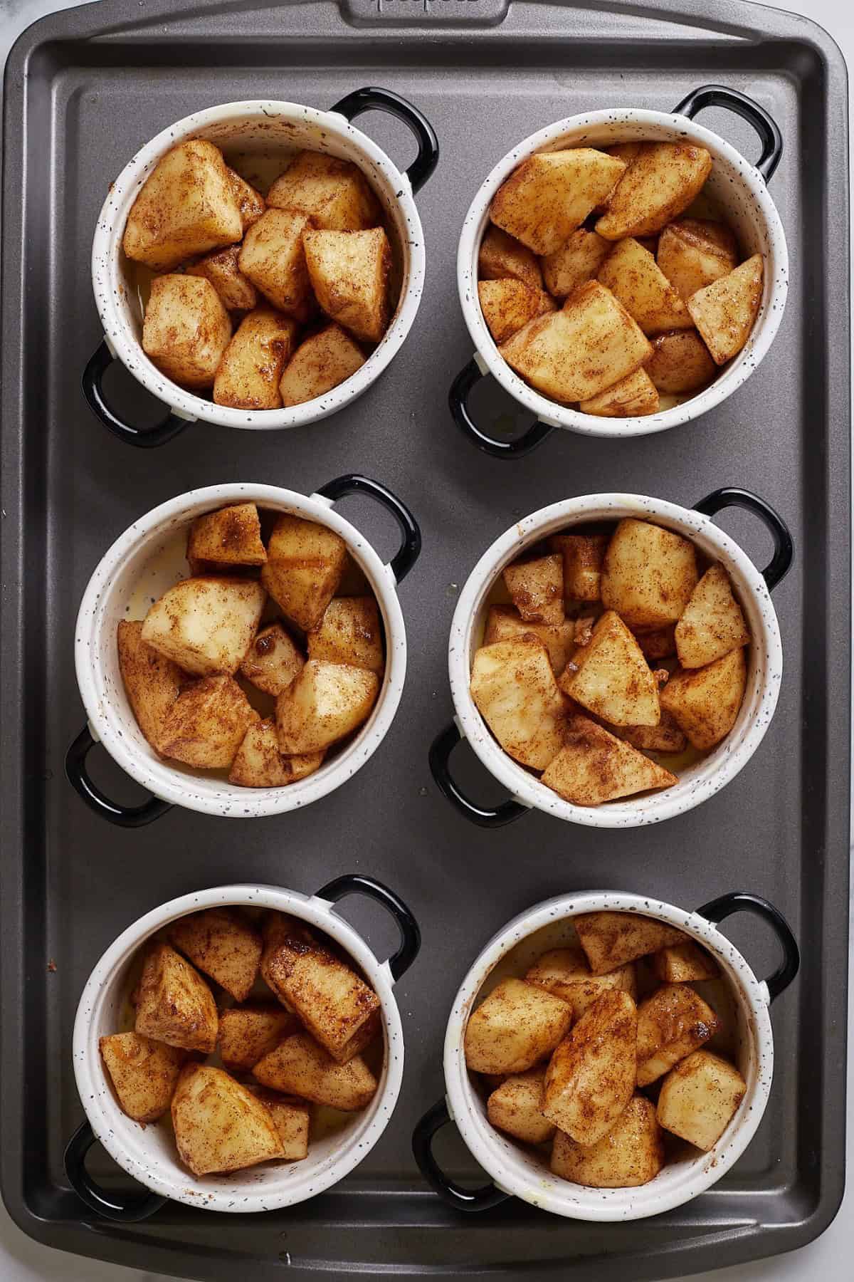 Seasoned apple mixture being evenly divided into six ramekins on a baking sheet.