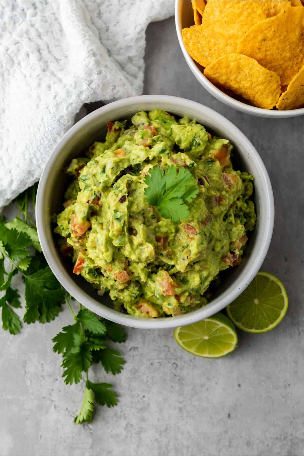A bowl of creamy guacamole surrounded by parsley leaves, a slice of lime, and nachos on the side.
