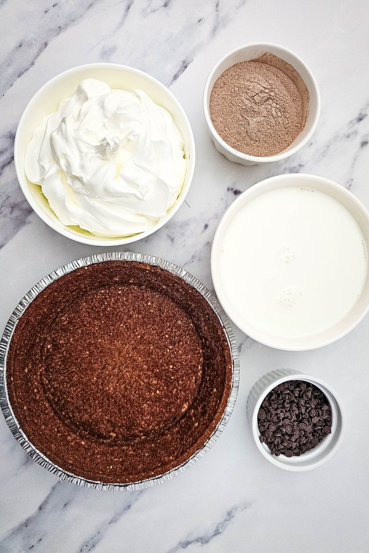 Chocolate pudding pie ingredients arranged on a surface, including pudding mix, milk, whipped topping, pie crust, and mini chocolate chips.