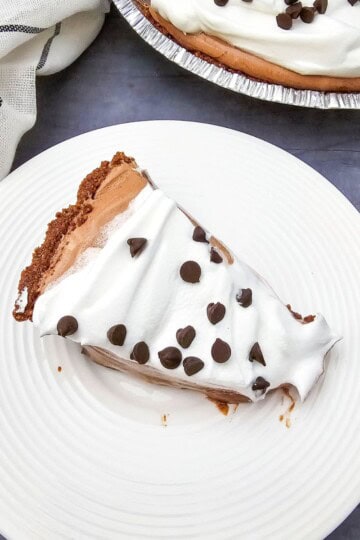 A slice of chocolate pie with whipped cream and chocolate chips on a plate, with the whole pie in the background.