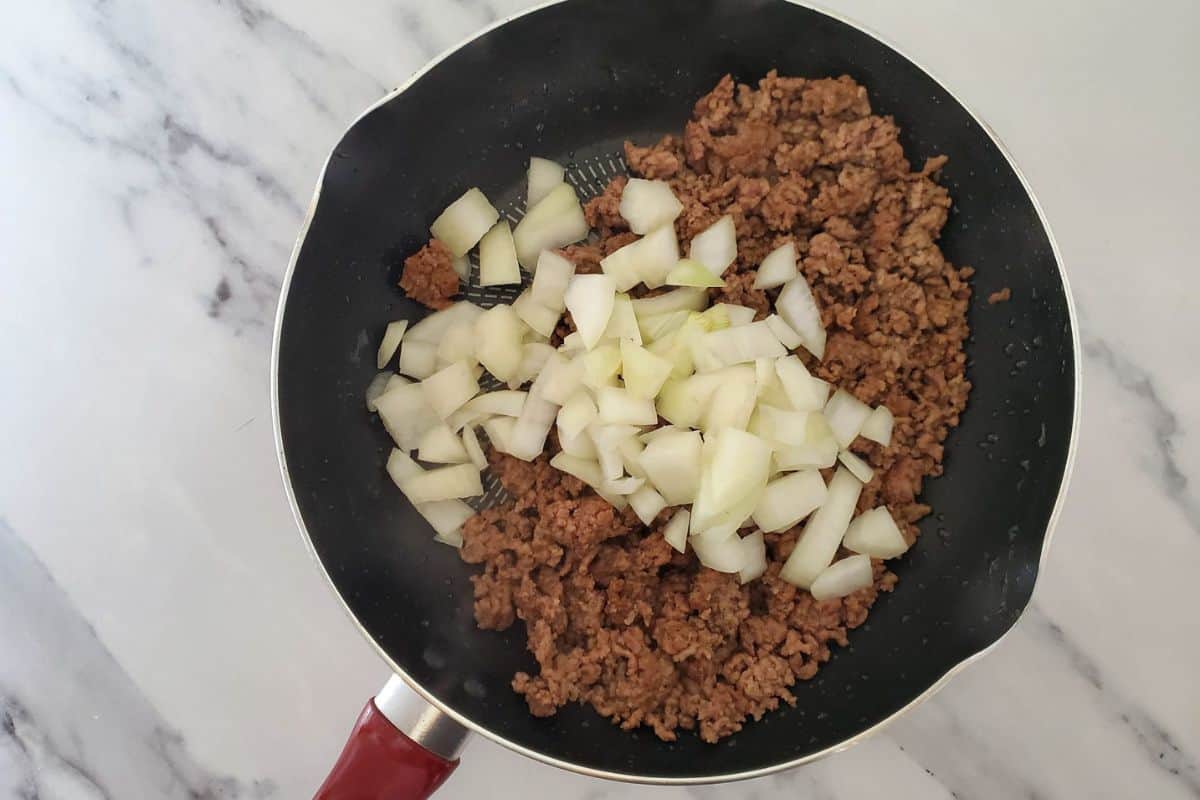 Ground beef sizzling in a pan, browning alongside diced onions and minced garlic.