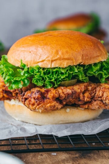 Close-up of a crispy chicken sandwich resting on a cooling rack, with more sandwiches slightly blurred in the background.