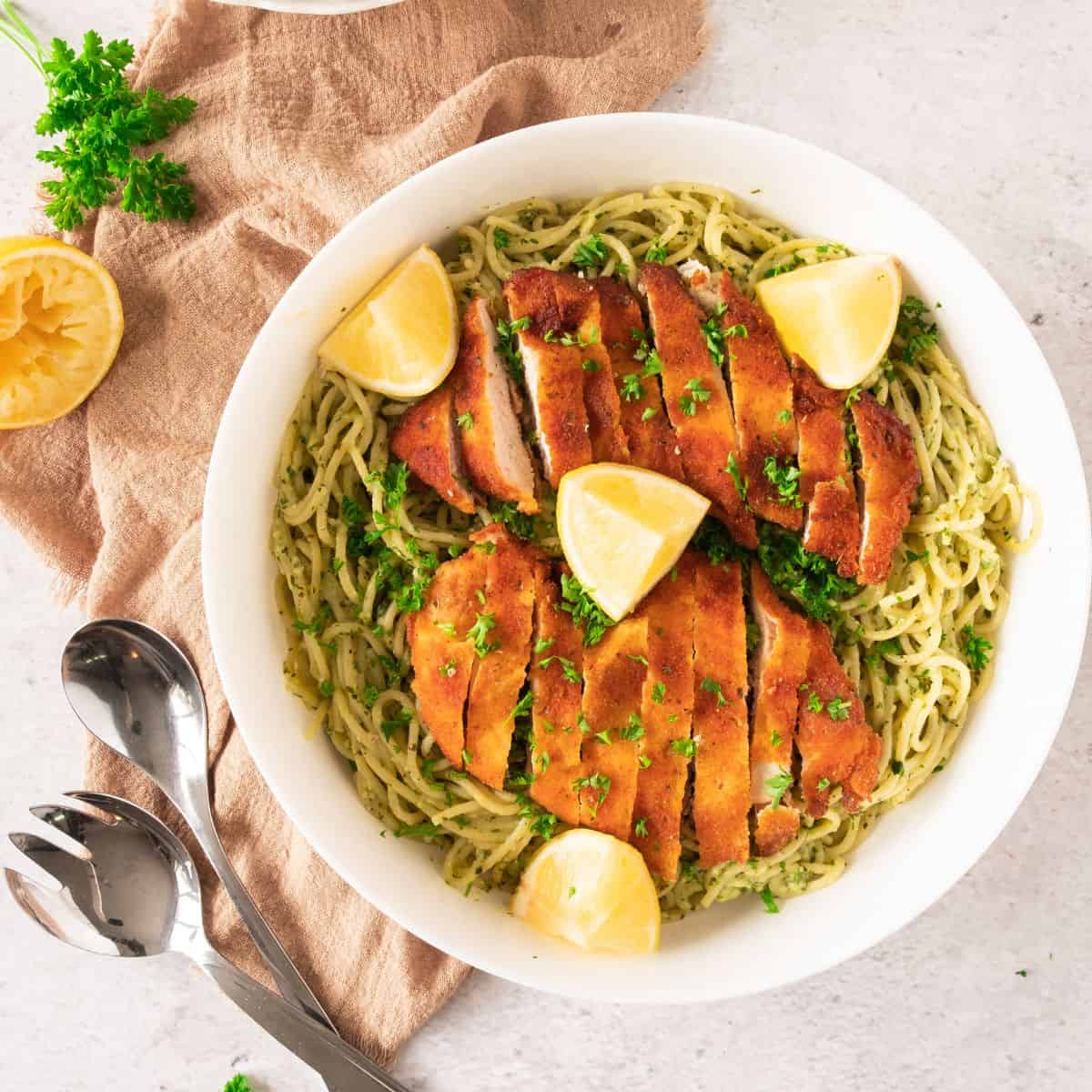 Crispy chicken over spaghetti in a bowl, topped with parsley and lemon, with a cloth underneath the bowl.