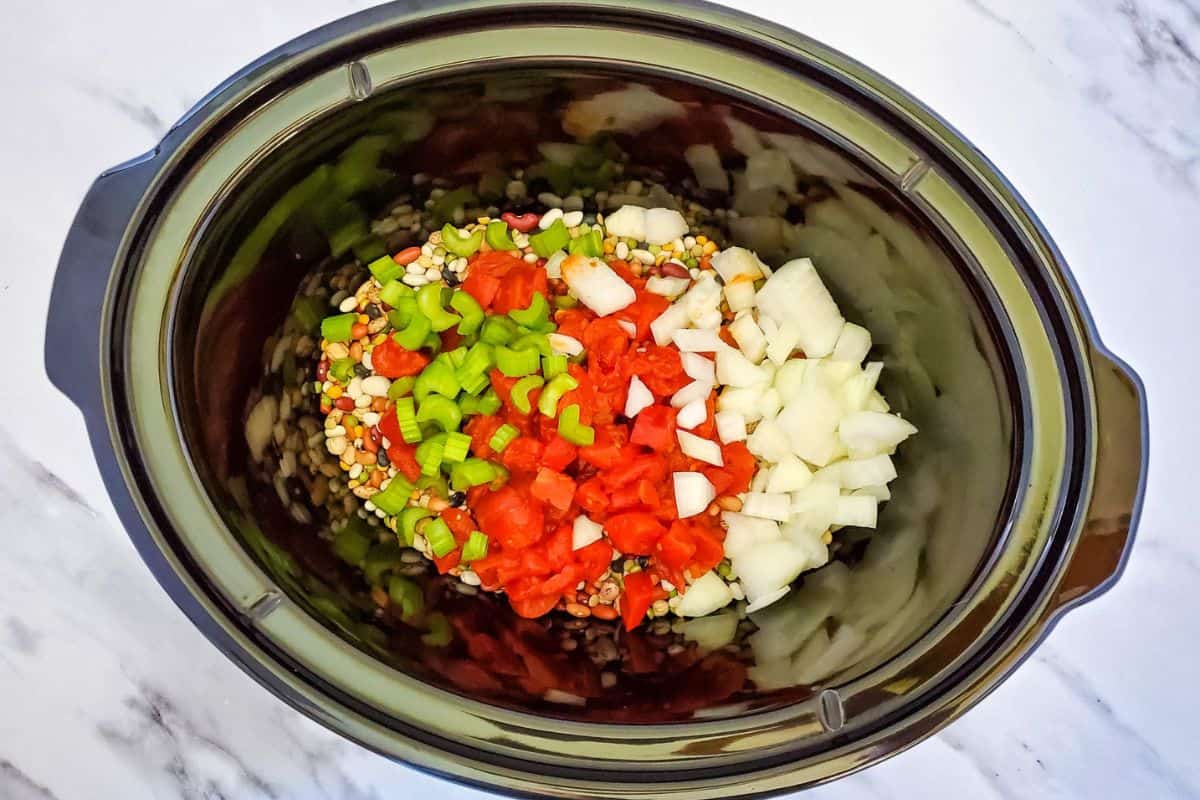 Ingredients being placed in a crockpot, including beans, chopped vegetables, and canned tomatoes.