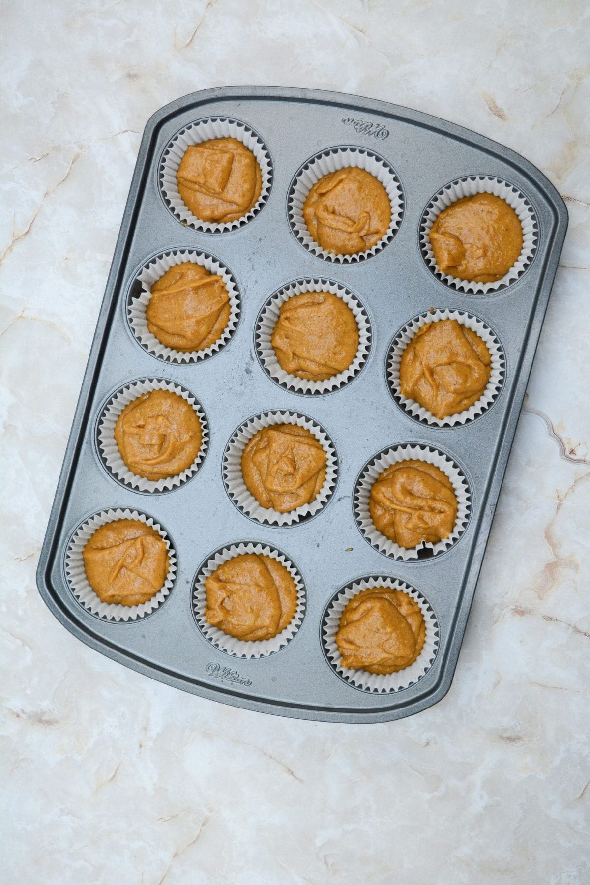Pumpkin muffin batter being spooned into paper-lined muffin cups in a baking tray, ready to be topped and baked.