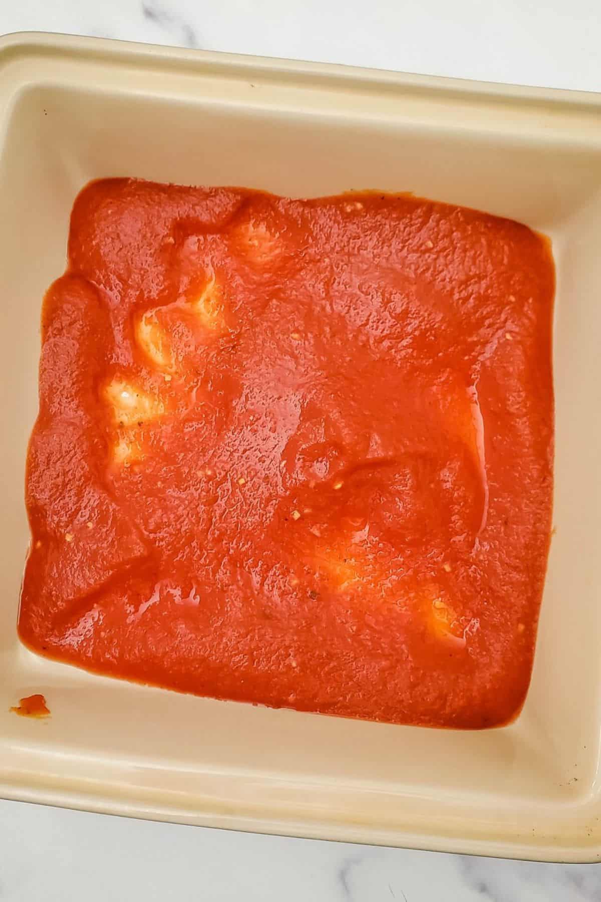 Ravioli in a container being topped with red pasta sauce.