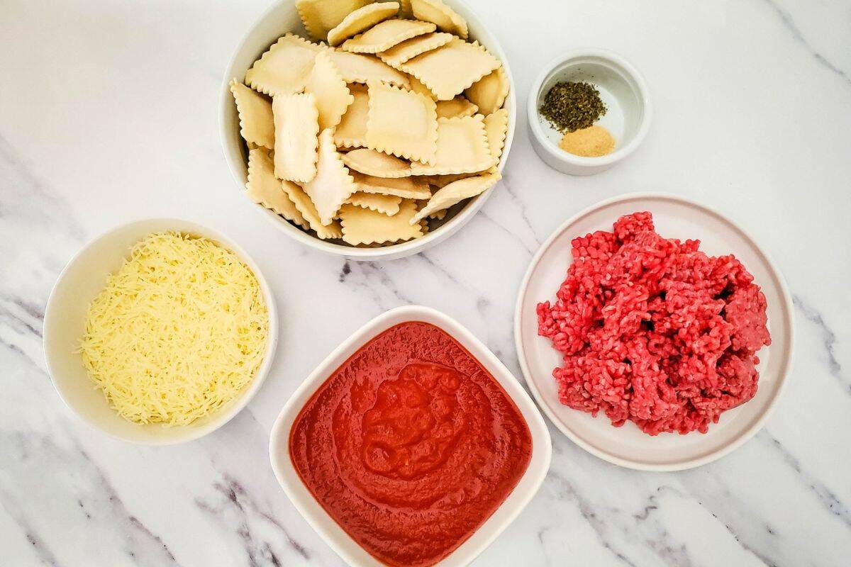 Ravioli lasagna ingredients laid out on a surface, including frozen ravioli, ground beef, pasta sauce, mozzarella cheese, and seasonings.