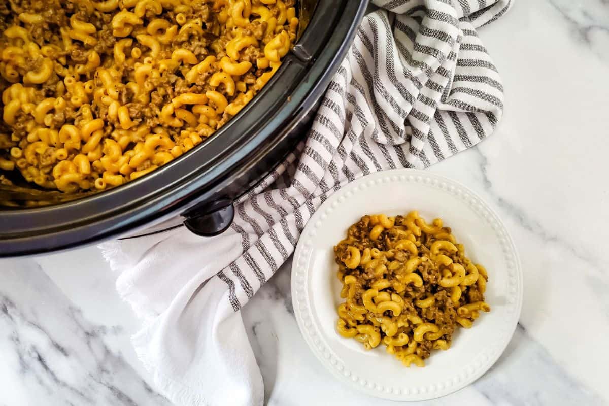 A plate of cheeseburger macaroni served beside a crockpot filled with more macaroni, partially covered with a kitchen cloth.