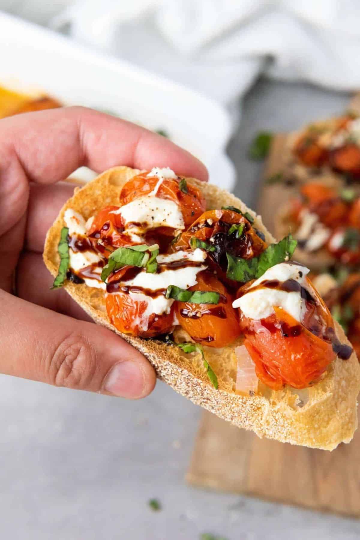 A person holding a roasted cherry tomato and burrata bruschetta, showing the creamy cheese and juicy tomatoes on toasted bread.
