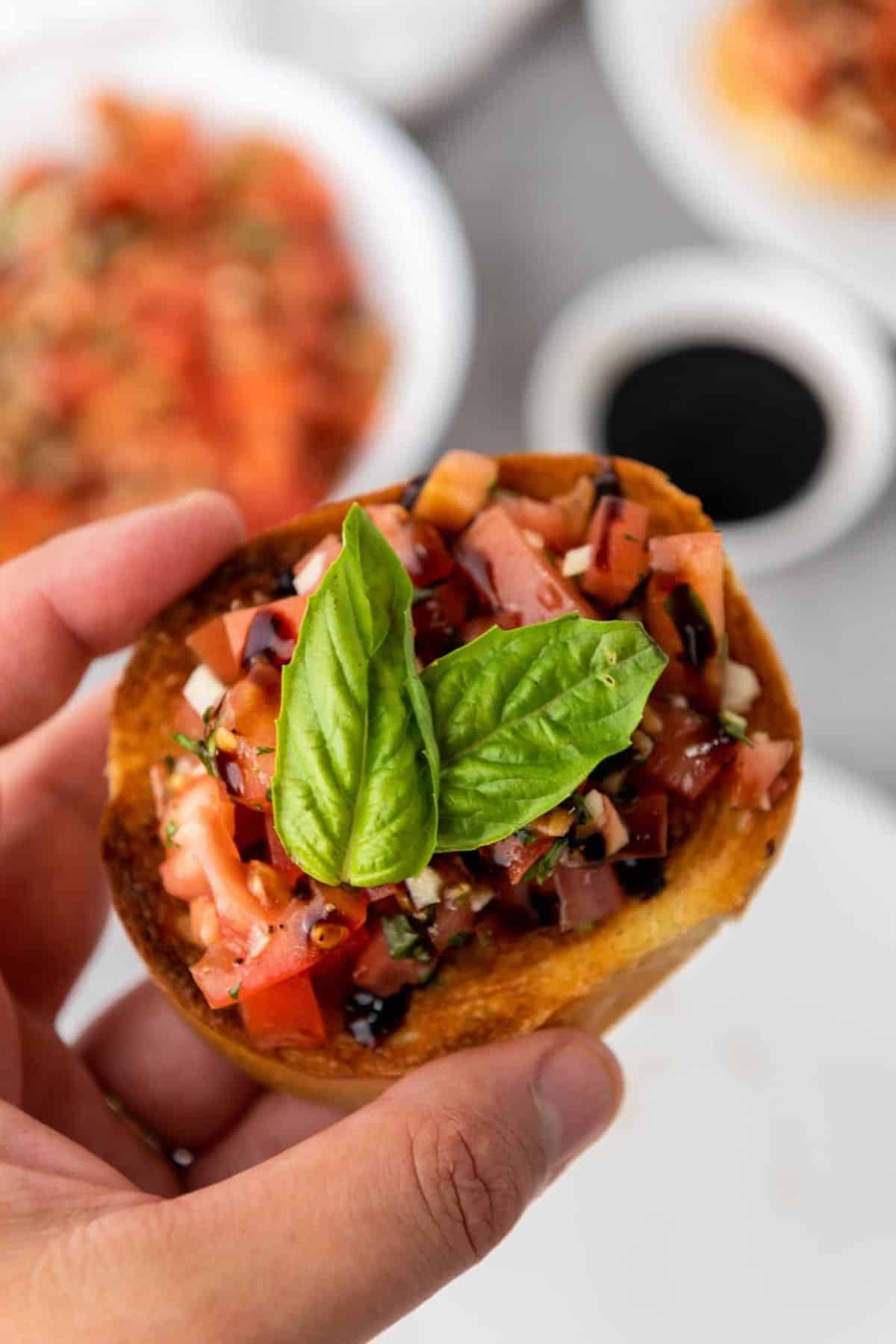A hand holding a single Classic Mediterranean Bruschetta topped with tomatoes and basil.
