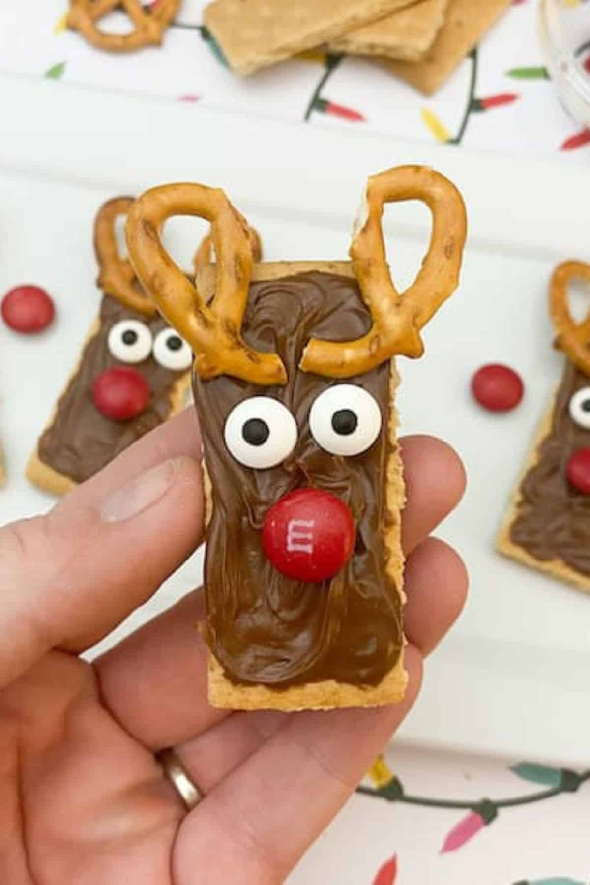 Hand holding a single Nutella Reindeer Cookie with pretzel antlers, with more cookies blurred in the background.