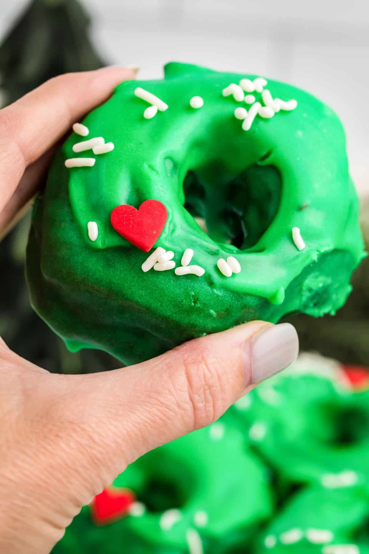 Hand holding a single green-frosted donut with sprinkles, with more donuts blurred in the blue background.