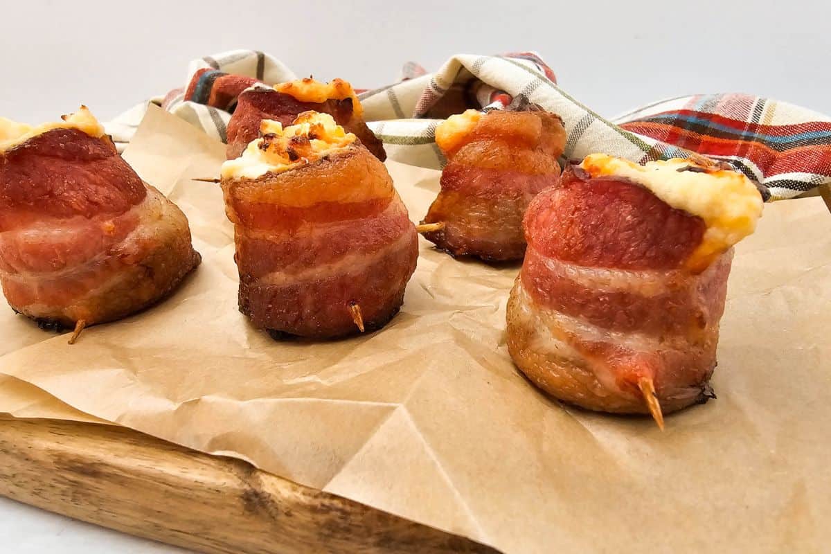 Close-up photo a bacon-wrapped pig shot filled with cheese served on a wooden board lined with parchment paper.