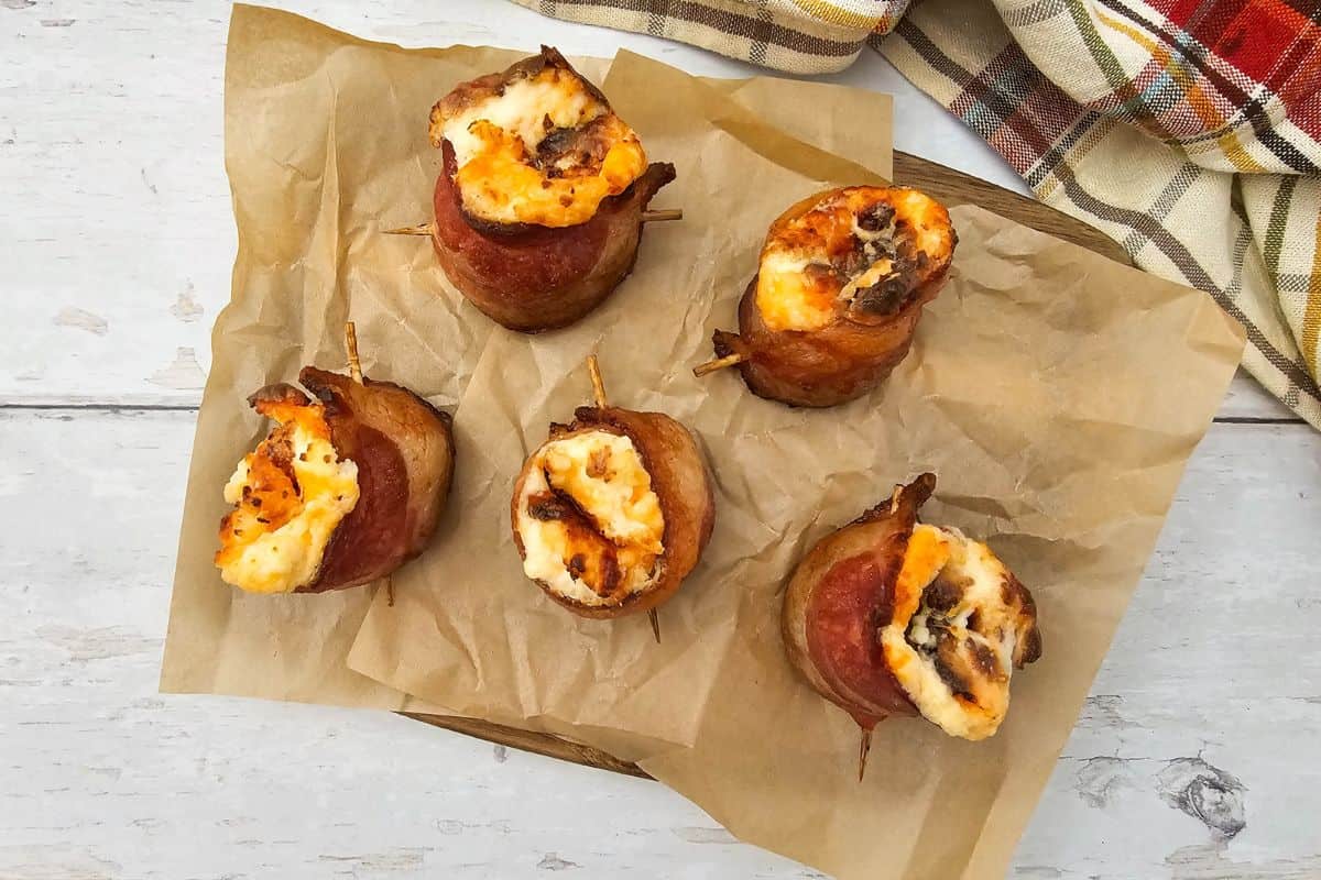 A bacon-wrapped pig shot filled with cheese served on a wooden board lined with parchment paper.