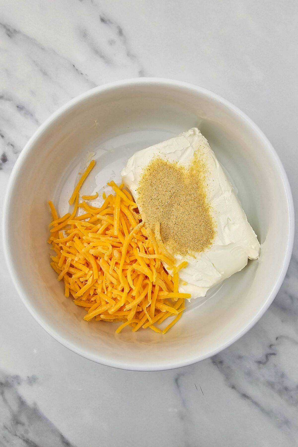 Ingredients including cream cheese, cheddar cheese and garlic powder being mixed in a bowl.