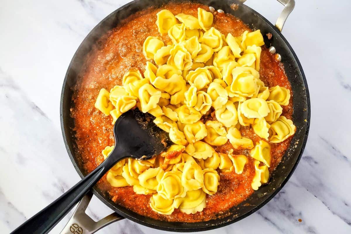 Pasta sauce simmering in a pot as cooked tortellini is added with a spatula.