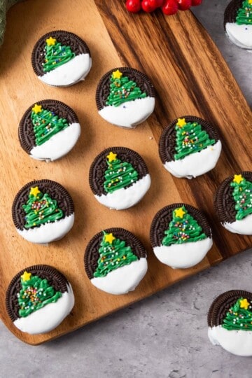 Christmas tree Oreos on a wooden board, placed on a cloth and surrounded by Christmas decorations.