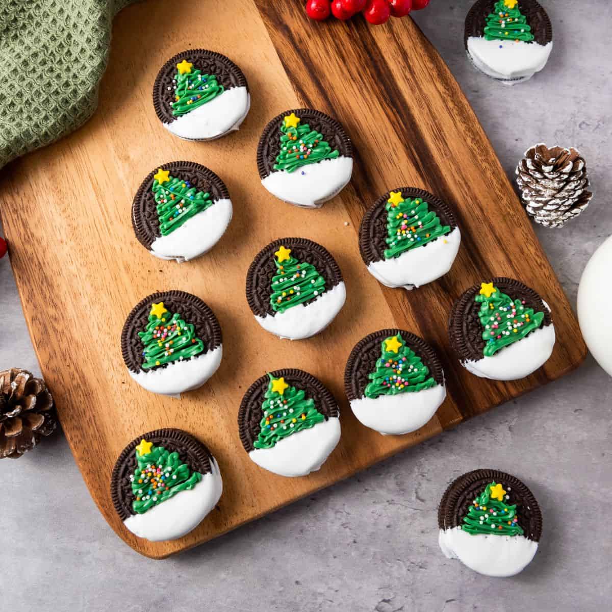Christmas tree Oreos on a wooden board, placed on a cloth and surrounded by Christmas decorations.