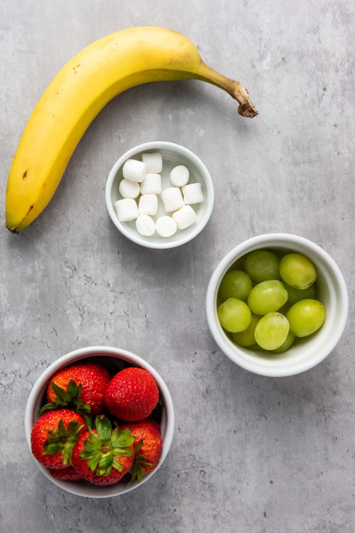 Ingredients for Green Christmas Man fruit kabobs, including green grapes, strawberries, and banana slices, arranged on a surface.