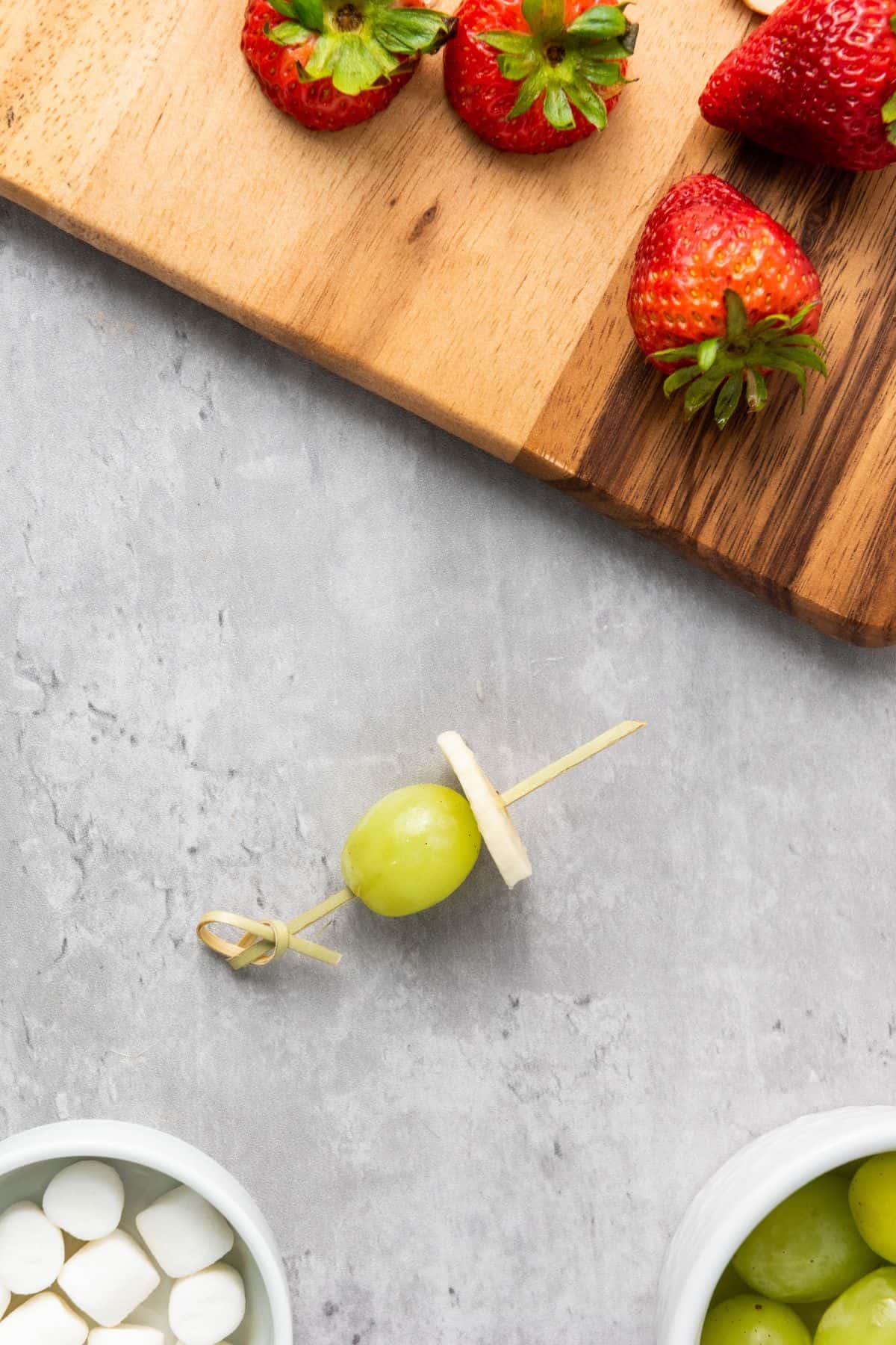 A grape and banana being threaded with strawberries on a wooden board and other ingredients surrounded by.