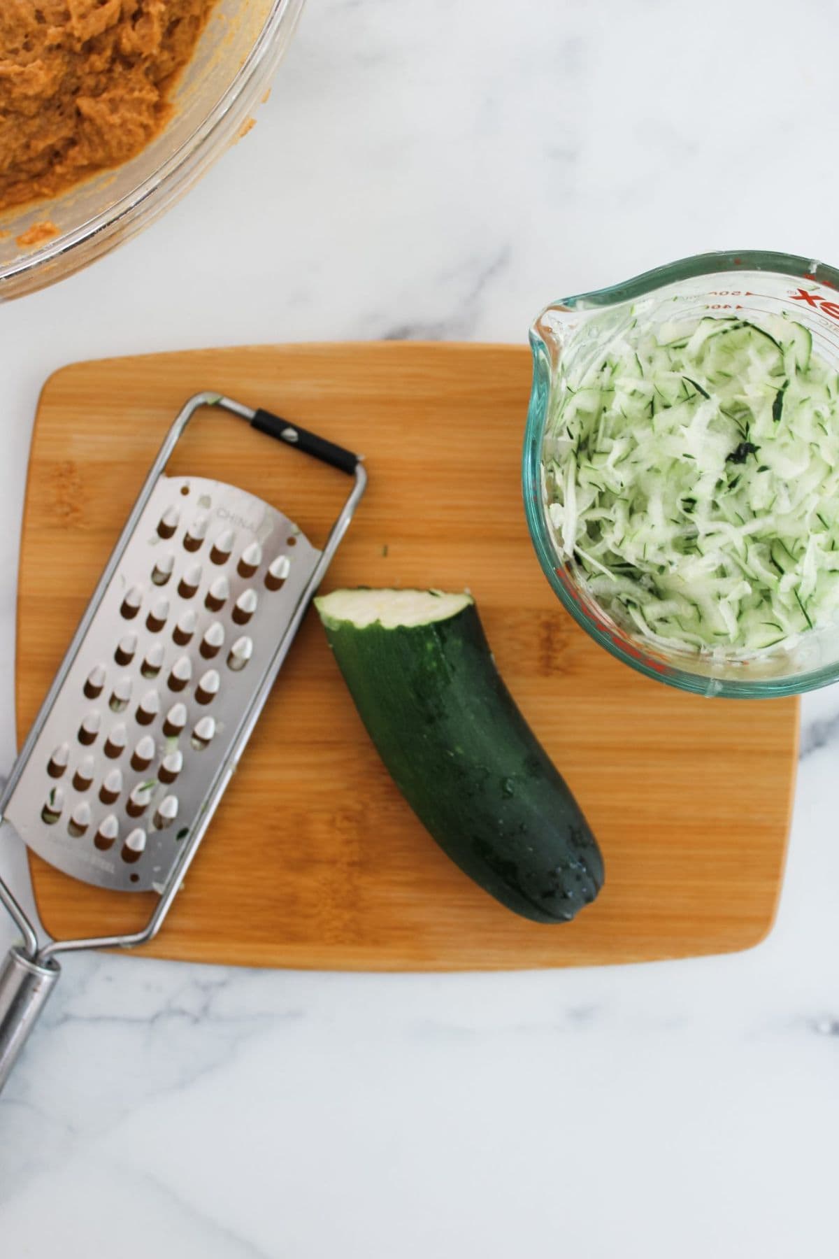 Half a zucchini and shredded pieces in a bowl, with a grater on a wooden board.