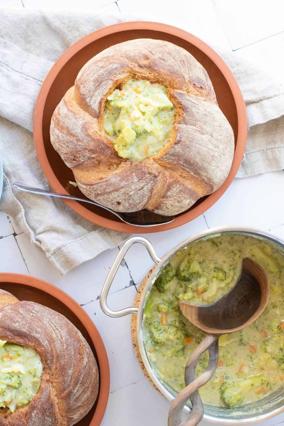 Broccoli cheddar soup served in a hollowed-out sourdough bread with a spoon on the side, alongside a pot filled with creamy soup and broccoli florets.