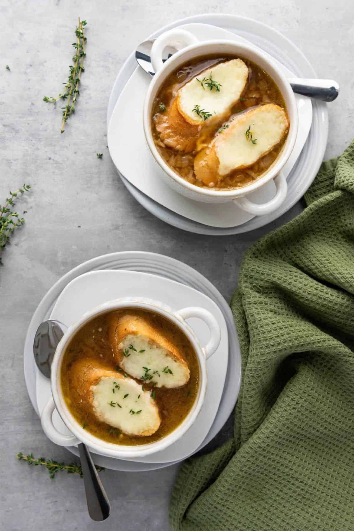 Two bowls of French Onion Soup, each topped with melted Gruyère over garlic bread, with a green cloth placed beside them.