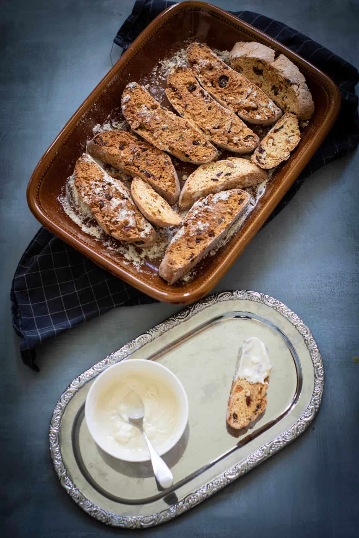 Biscotti log being sliced with a serrated knife in a baking dish, with a small bowl of melted white chocolate and a single biscotti dipped in it.