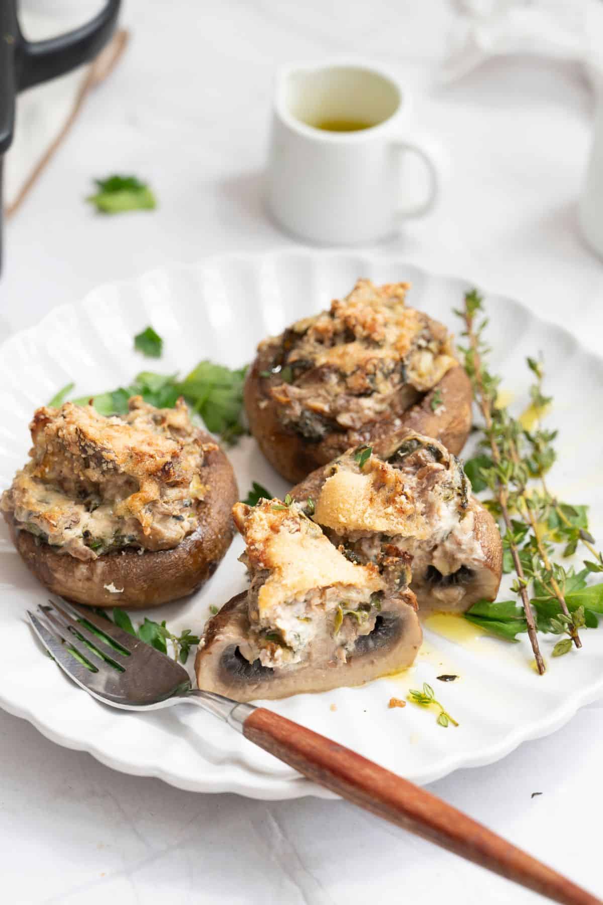 Stuffed mushrooms with spinach & marinated artichokes on a plate, one mushroom cut open to show the creamy filling, with a fork beside it and garnished with fresh herbs.
