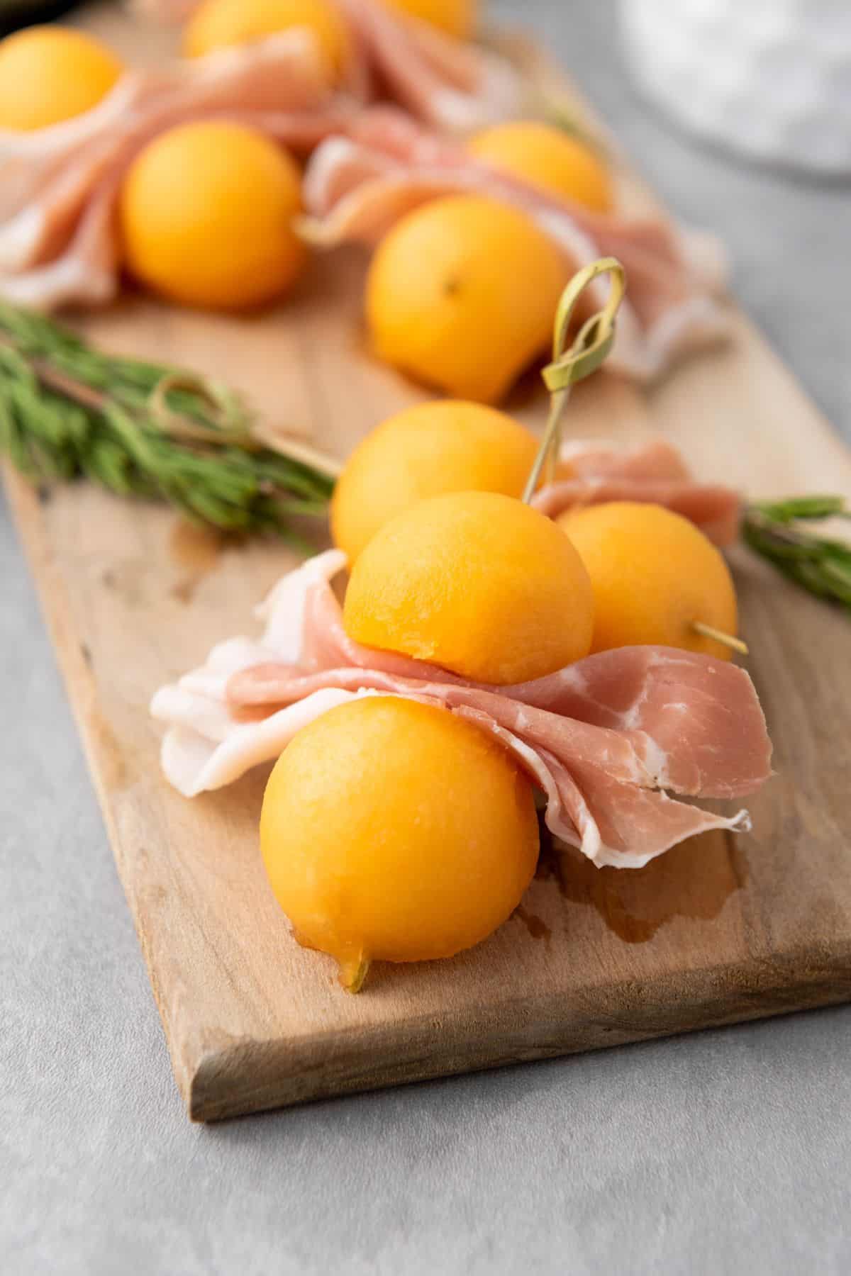Cantaloupe prosciutto skewers resting on a wooden board, garnished with fresh herbs.