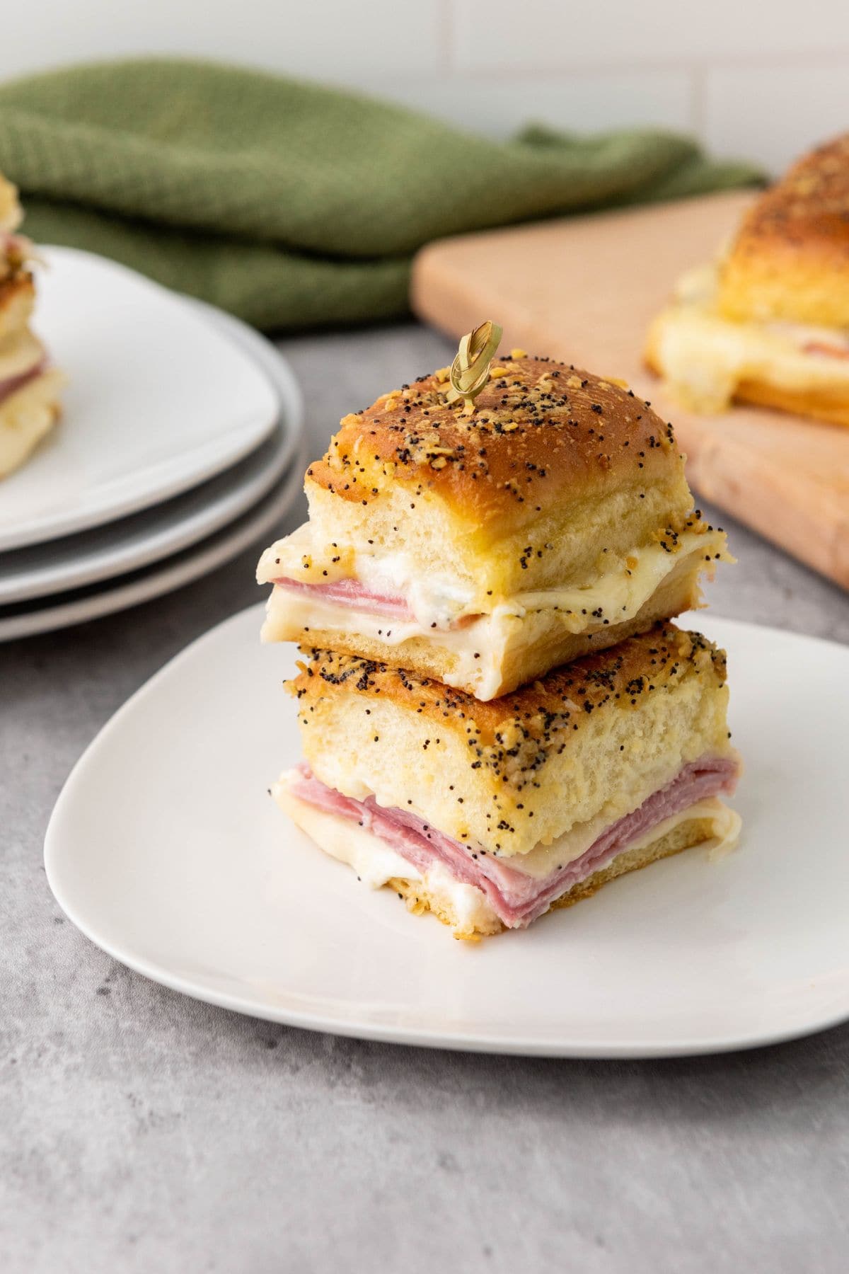 Melty Ham and Cheese Sliders stacked on a plate with the background showing blurred Hawaiian rolls.