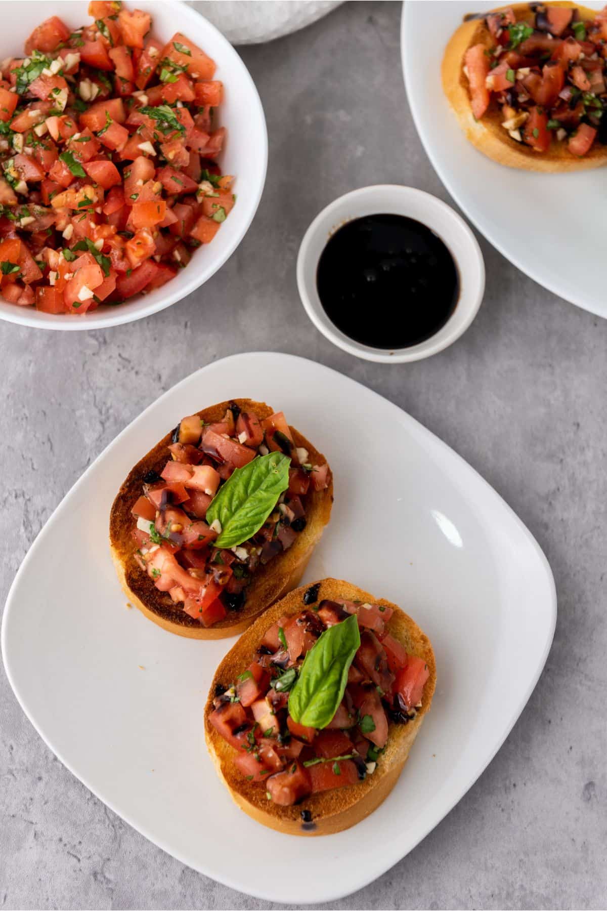 Two pieces of classic Mediterranean bruschetta on a plate, with extra tomato and basil filling in a small bowl and balsamic glaze on the side.