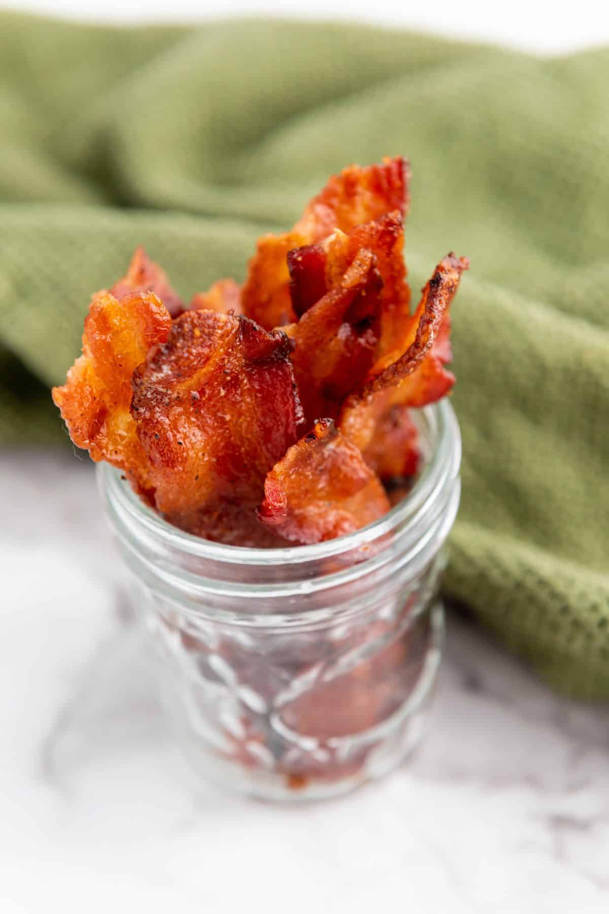 Candied maple brown sugar bacon stacked in a small jar with a green cloth beside it.