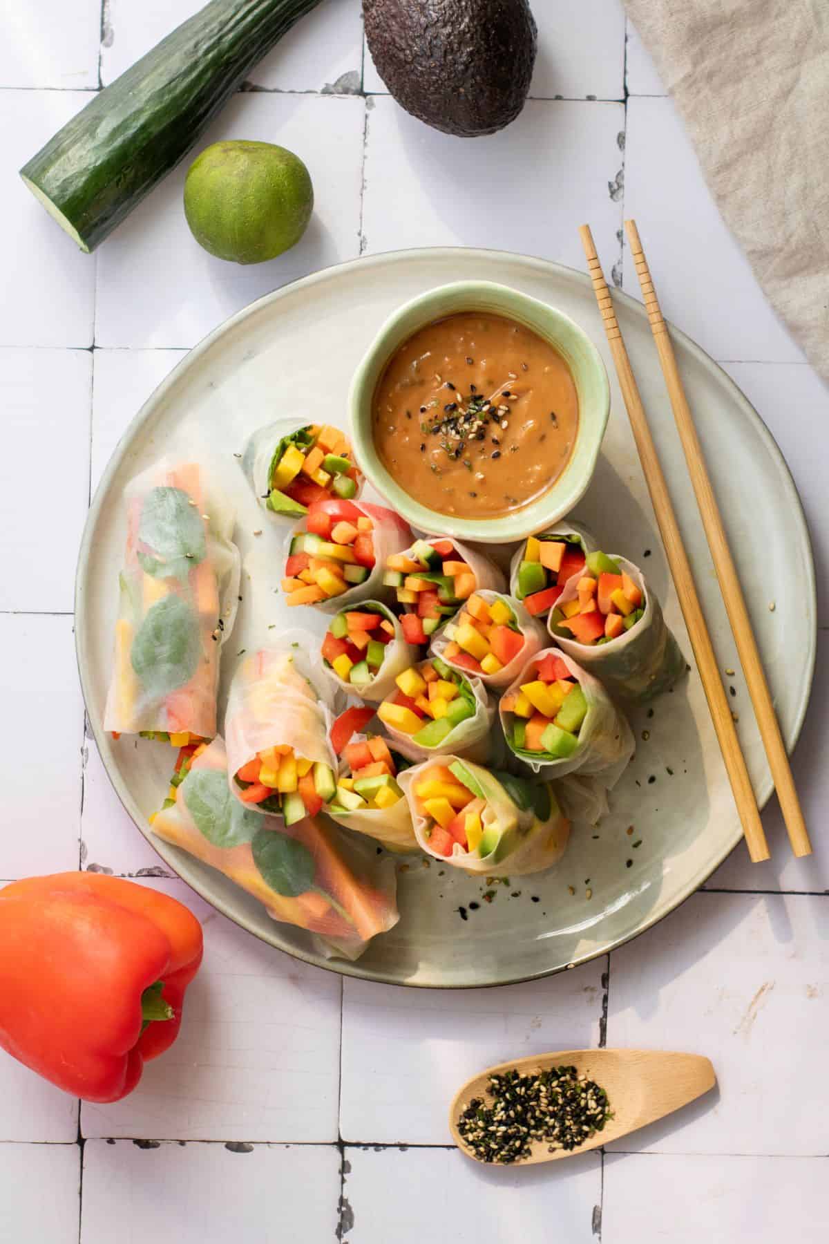 A plate filled with rainbow veggie rice paper rolls, served with a small bowl of dipping sauce and chopsticks on the side.