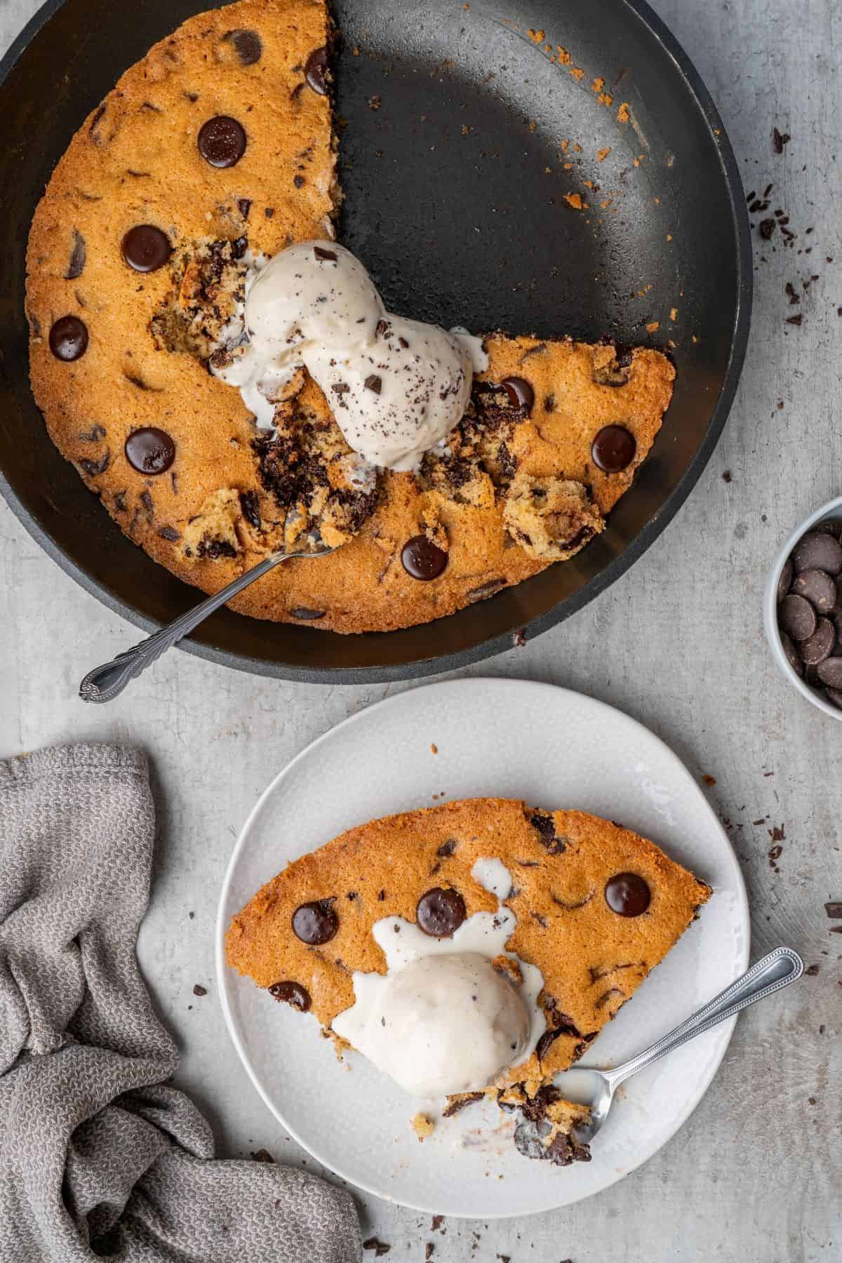 Chocolate chip skillet cookies topped with vanilla ice cream in a skillet, with a plate filled with a serving beside it.